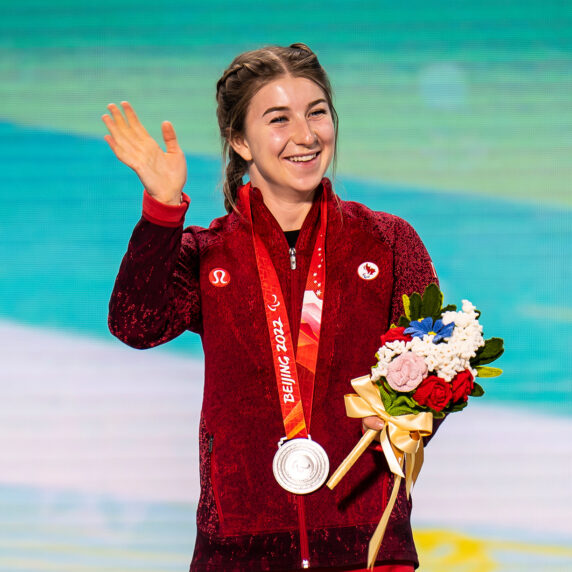 La paralympienne canadienne Mollie Jepsen se tient sur le podium des Jeux paralympiques d’hiver de Beijing 2022, portant une veste rouge de l’équipe du Canada. Elle salue et sourit tout en tenant sa médaille d’argent et un bouquet de fleurs, célébrant son exploit en ski para-alpin.