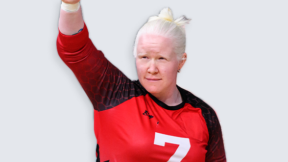 La Paralympienne canadienne Amy Burk, joueuse de goalball, portant un maillot rouge et noir d'Équipe Canada avec le numéro 7. Elle lève son bras droit avec une expression concentrée, ses cheveux blond platine attachés en chignon haut.