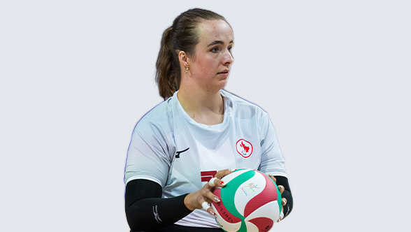 La Paralympienne canadienne Heidi Peters, joueuse de volleyball assis, portant un maillot blanc d'Équipe Canada avec des manches noires. Elle tient un ballon de volleyball rouge, blanc et vert à deux mains, prête à servir, avec une expression concentrée sur son visage.