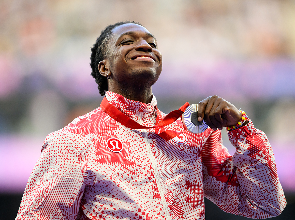 Le paralympien canadien Jesse Zesseu sourit fièrement en tenant sa médaille d’argent aux Jeux paralympiques de Paris 2024. Il porte la veste de podium rouge et blanche de l’équipe canadienne conçue par lululemon, arborant un motif abstrait audacieux. Les lumières floues du stade en arrière-plan soulignent ce moment de célébration.