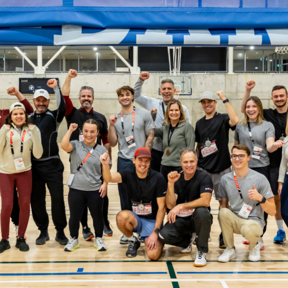 n groupe de participants à la ParaTough Cup de Montréal 2024 pose ensemble dans un gymnase, souriant et levant le poing en signe de célébration. Ils portent des vêtements de sport et des lanières d’événement, debout sur un terrain en bois avec un panier de basketball et des bannières bleues en arrière-plan.