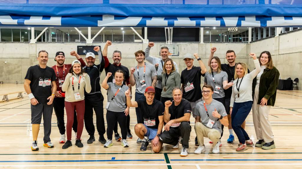 n groupe de participants à la ParaTough Cup de Montréal 2024 pose ensemble dans un gymnase, souriant et levant le poing en signe de célébration. Ils portent des vêtements de sport et des lanières d’événement, debout sur un terrain en bois avec un panier de basketball et des bannières bleues en arrière-plan.