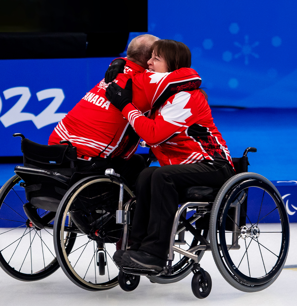 Deux curleurs en fauteuil roulant de l’équipe canadienne s’étreignent en célébration sur la glace. Ils portent des vestes rouges et blanches assorties avec des feuilles d’érable. Les deux athlètes sont en fauteuil roulant, et l’arrière-plan présente une patinoire bleue avec les chiffres « 2022 », indiquant les Jeux paralympiques d’hiver de Beijing 2022.