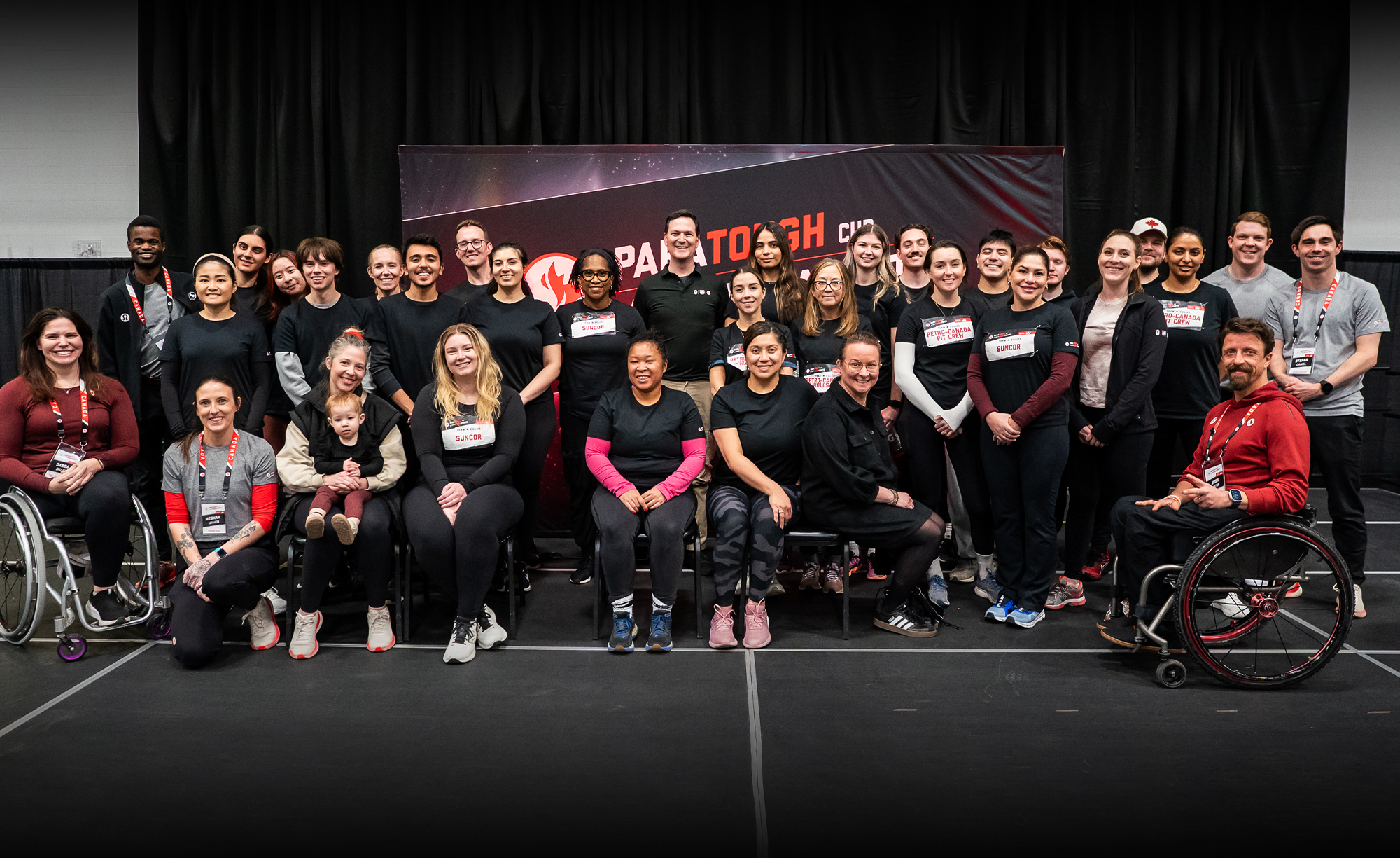 Une photo de groupe du personnel de Petro-Canada et des paralympiens canadiens lors de l'événement ParaTough Cup à Calgary. Les participants sont rassemblés devant une toile de fond noire et rouge avec le logo ParaTough Cup, souriant et posant ensemble. Certaines personnes sont assises, y compris des athlètes en fauteuil roulant, tandis que d'autres se tiennent debout derrière eux. Le groupe reflète une diversité d'âges, d'origines et de rôles, uni dans son soutien au para sport.