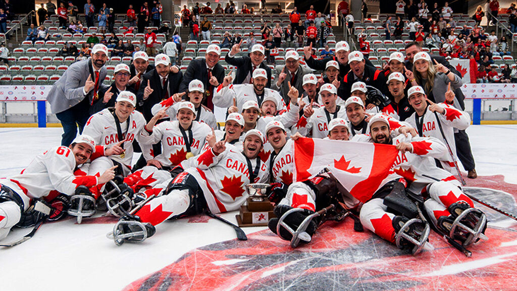 The Canadian National Para Hockey Team celebrates on the ice after winning gold at the 2024 World Para Hockey World Championships. The players, dressed in white Team Canada jerseys with red maple leaf accents, pose with the championship trophy, waving a Canadian flag and making number-one gestures. Behind them, coaches and staff join in the celebration, with excited fans in the stands.