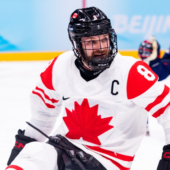 Le joueur canadien de parahockey Tyler McGregor, portant le "C" de capitaine sur son chandail blanc d’Équipe Canada orné d’une feuille d’érable rouge, participe à un match. Il est concentré sur l’action, tenant fermement ses bâtons de hockey tandis qu’un joueur des États-Unis apparaît en arrière-plan.