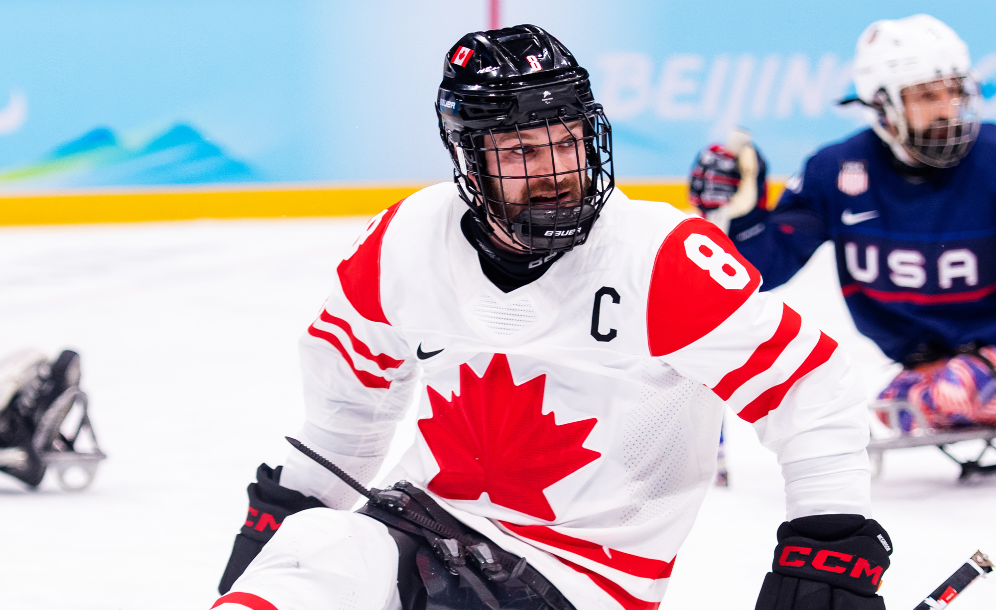 Le joueur canadien de parahockey Tyler McGregor, portant le "C" de capitaine sur son chandail blanc d’Équipe Canada orné d’une feuille d’érable rouge, participe à un match. Il est concentré sur l’action, tenant fermement ses bâtons de hockey tandis qu’un joueur des États-Unis apparaît en arrière-plan.