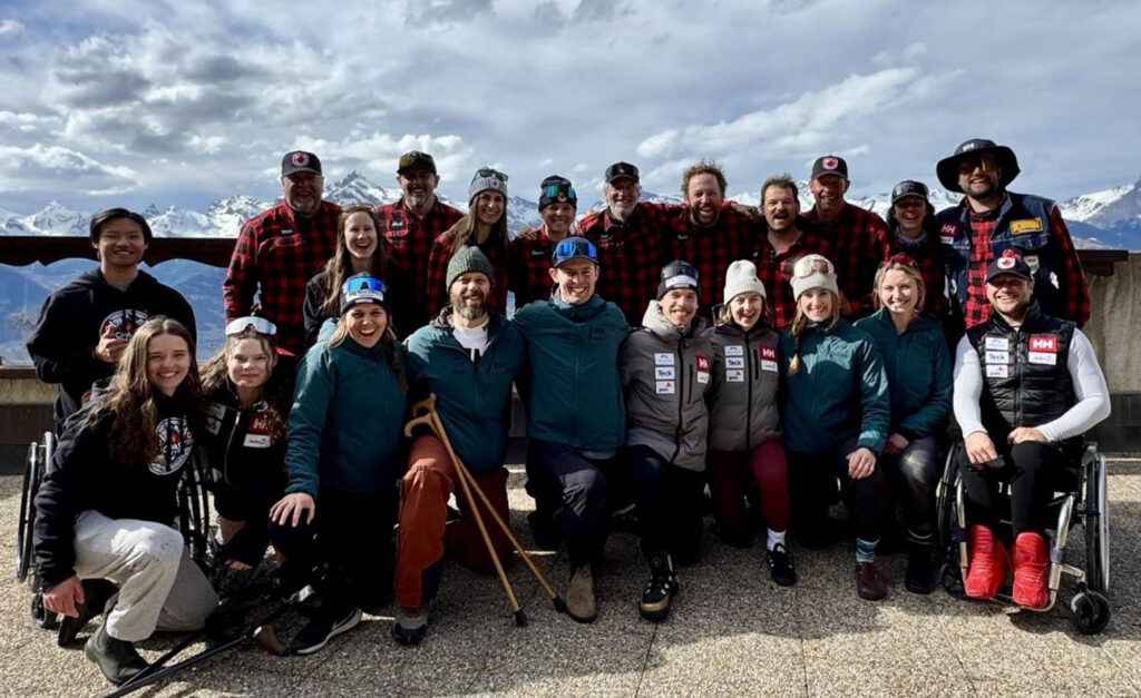 Photo de groupe de l’équipe canadienne de ski para-alpin souriant à l’extérieur, avec des montagnes enneigées en arrière-plan. Les membres de l’équipe portent un mélange de vestes à carreaux, de vêtements d’hiver et de tenues d’équipe.