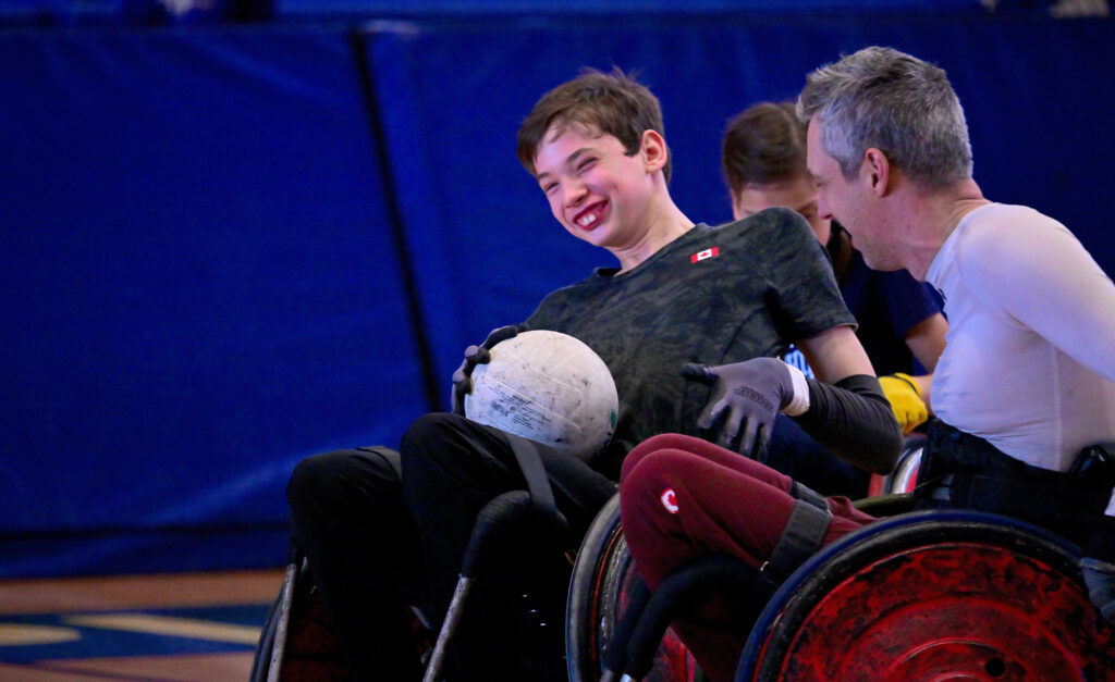 Un jeune joueur canadien de rugby en fauteuil roulant sourit de joie en tenant un ballon de rugby lors d’un match amical contre le paralympien canadien Byron Green. Les deux athlètes sont assis dans des fauteuils roulants sportifs sur un terrain de gymnase, partageant un moment de complicité et de plaisir.