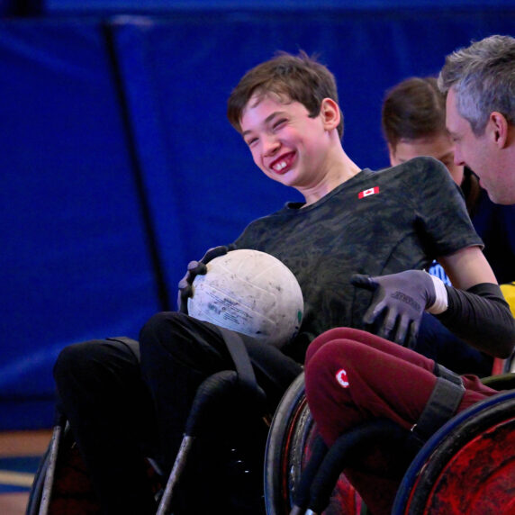 Un jeune joueur canadien de rugby en fauteuil roulant sourit de joie en tenant un ballon de rugby lors d’un match amical contre le paralympien canadien Byron Green. Les deux athlètes sont assis dans des fauteuils roulants sportifs sur un terrain de gymnase, partageant un moment de complicité et de plaisir.