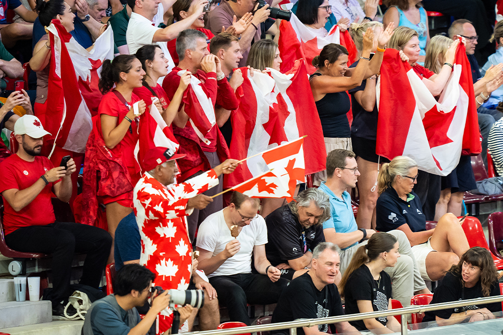 Canadian fans cheer loudly from the stands at the Paris 2024 Paralympic Games, waving Canadian flags and dressed in red and white. One fan stands out in a full maple leaf suit, proudly holding two flags, as the crowd shows their enthusiastic support for the Canadian Paralympic Team.