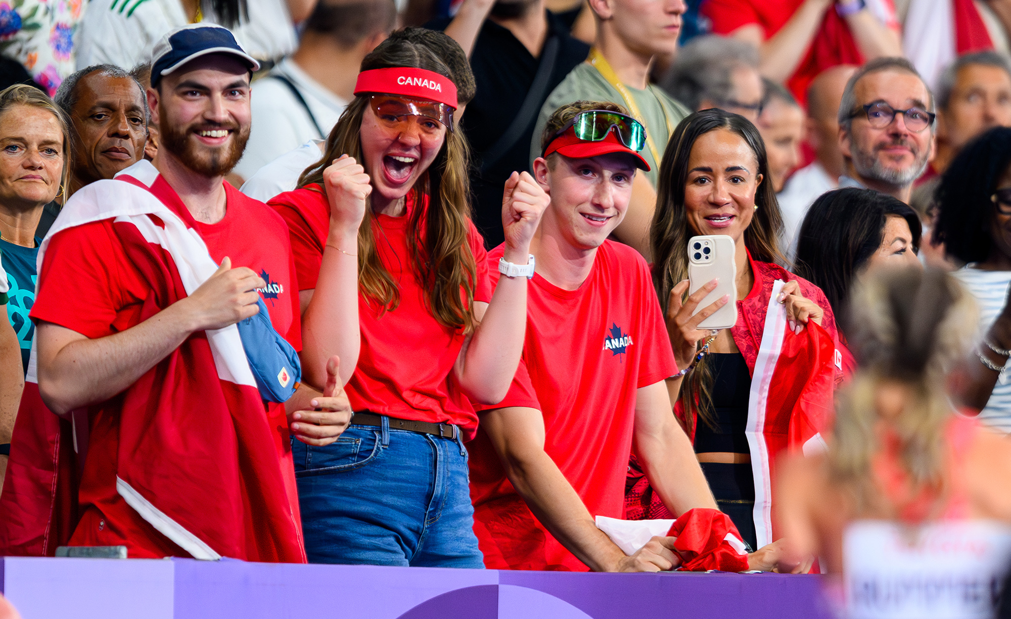 Un groupe de fervents partisans canadiens, vêtus de rouge et agitant des drapeaux du Canada, encourage bruyamment la paralympienne canadienne Amanda Rummery aux Jeux paralympiques de Paris 2024. Un fan immortalise l’instant sur son téléphone, tandis que d’autres lèvent les poings avec enthousiasme, exprimant leur fierté et leur soutien.