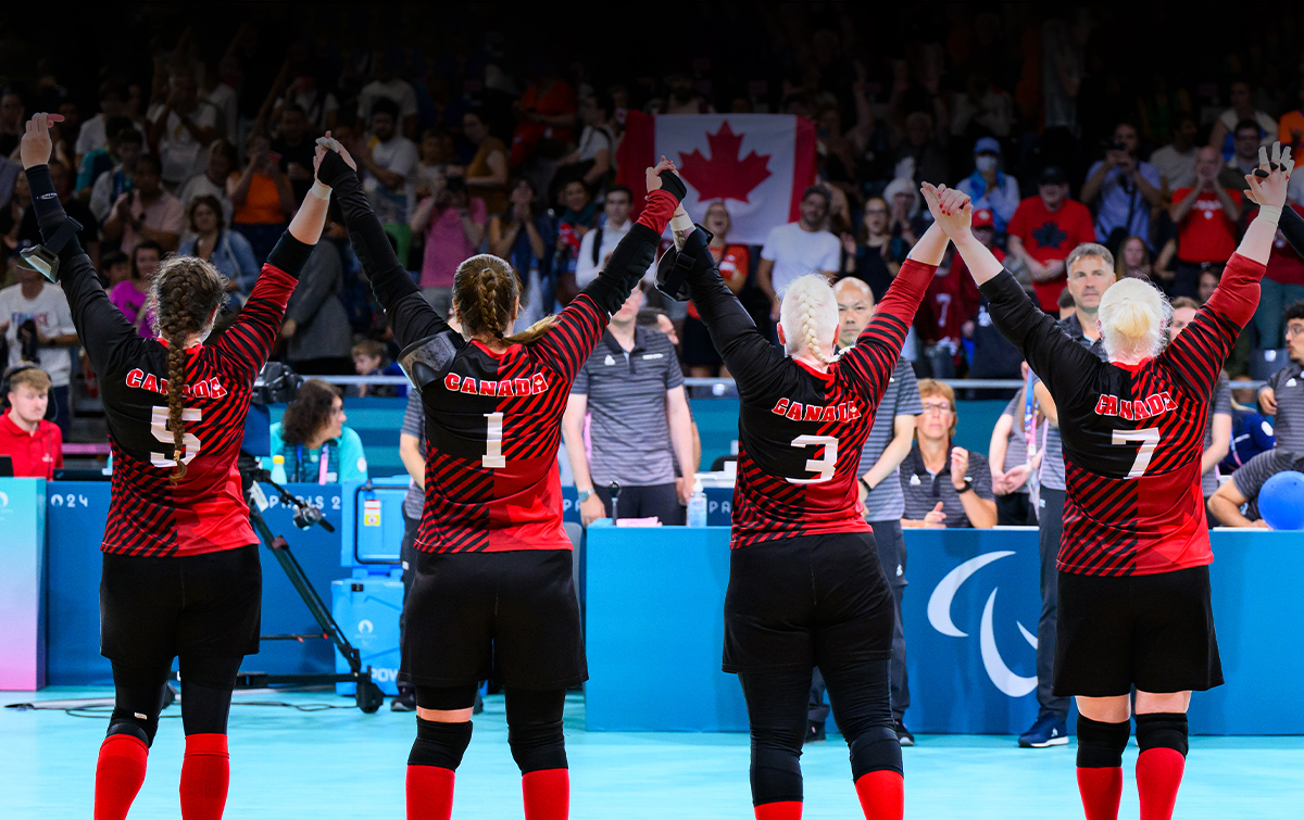 L’équipe canadienne féminine de goalball célèbre en levant les bras, face à la foule, après un match aux Jeux paralympiques de Paris 2024. Les partisans applaudissent et agitent un drapeau canadien en arrière-plan.