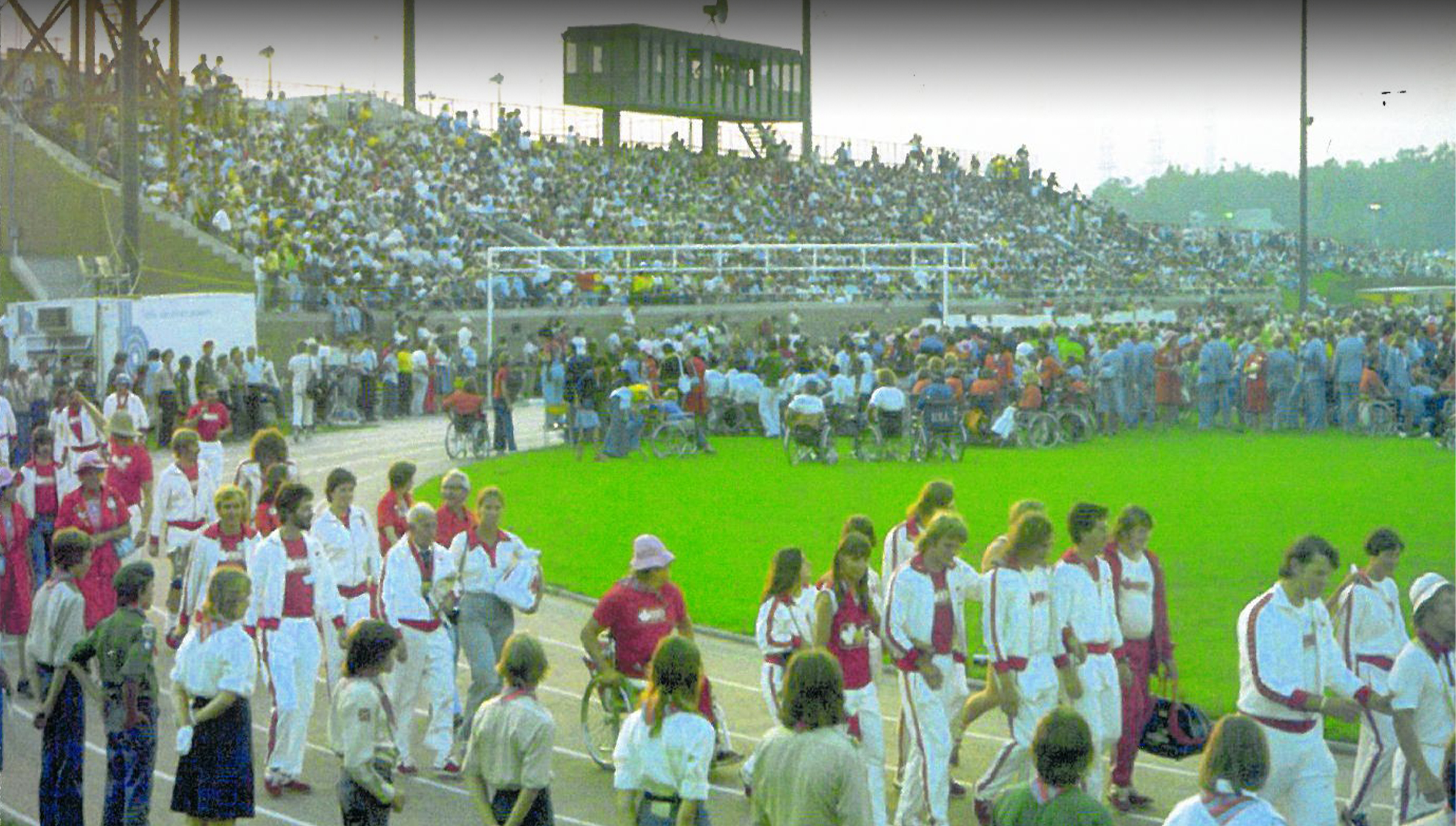 Des athlètes paralympiques défilent sur la piste du stade lors de la cérémonie d’ouverture des Jeux paralympiques de Toronto 1976, sous les yeux d’une foule nombreuse rassemblée dans les gradins.