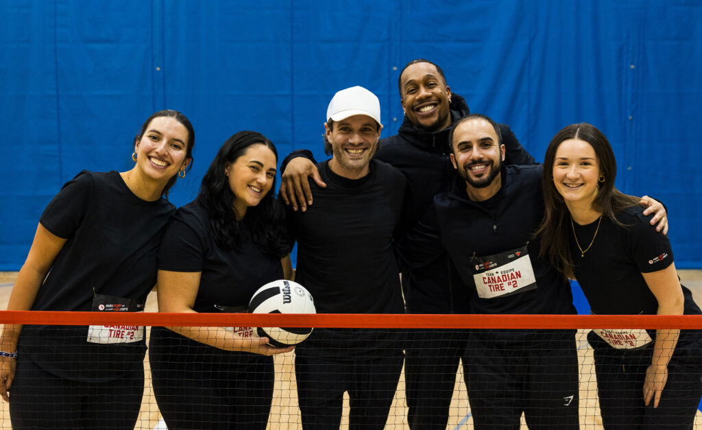 Une équipe souriante de Canadian Tire pose ensemble sur un terrain de gymnase derrière un filet lors de l'événement ParaTough Cup à Toronto, tenant un ballon de volleyball et vêtue de chandails noirs assortis.