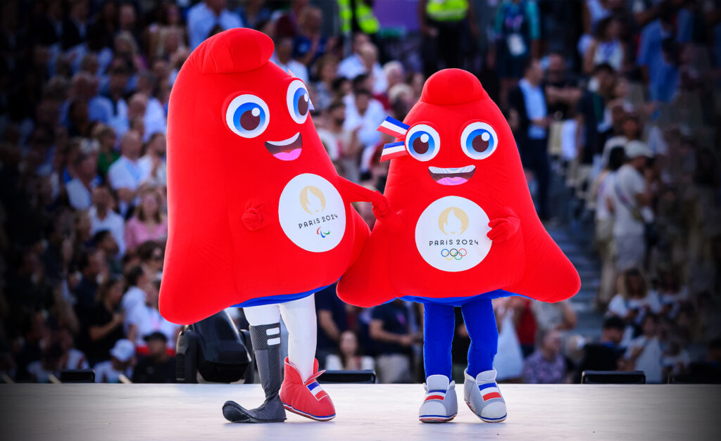 Les mascottes de Paris 2024, appelées les Phryges, apparaissent sur scène lors d’un événement public. Les personnages rouges en forme de bonnet sourient et se tiennent la main, l’un représentant les Jeux paralympiques et l’autre les Jeux olympiques, chacun arborant l’emblème de Paris 2024 sur leur torse.