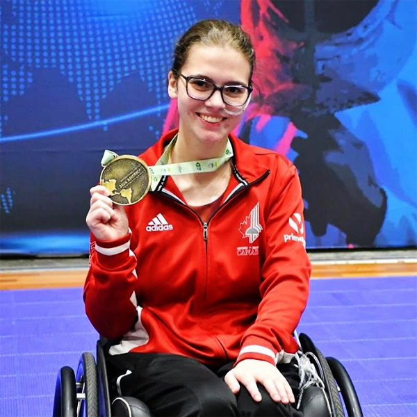Canadian wheelchair fencer Trinity Lowthian smiles while holding up a gold medal. She is wearing a red Team Canada jacket and sitting in her wheelchair on a competition floor.