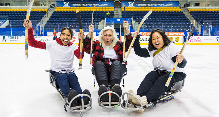 Three enthusiastic ParaTough Cup volunteers smiling and cheering while seated on sleds on the ice, holding sledge hockey sticks inside an arena.