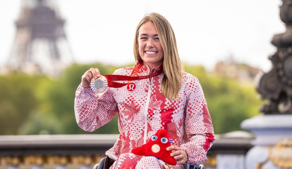 La paralympienne canadienne Leanne Taylor sourit en tenant sa médaille de bronze de Paris 2024 et la peluche mascotte paralympique. Elle porte une veste de podium rouge et blanche d’Équipe Canada, avec la tour Eiffel floue en arrière-plan.