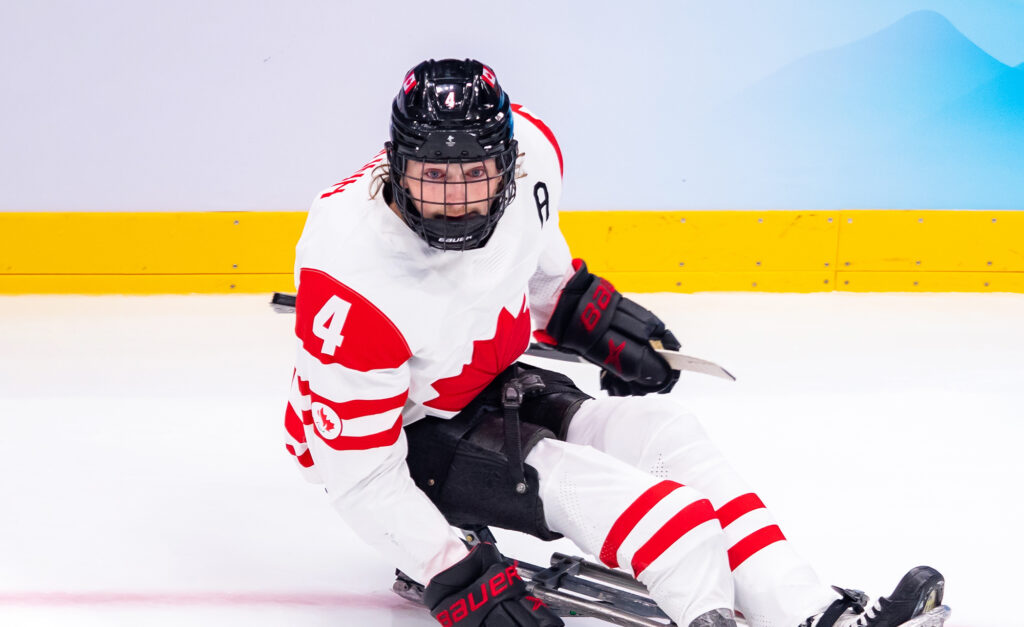 Le joueur canadien de hockey sur luge James Dunn, portant le numéro 4, est en action sur la glace pendant un match. Il porte l’uniforme blanc de l’équipe du Canada avec des accents rouges et un casque noir, concentré sur le jeu.