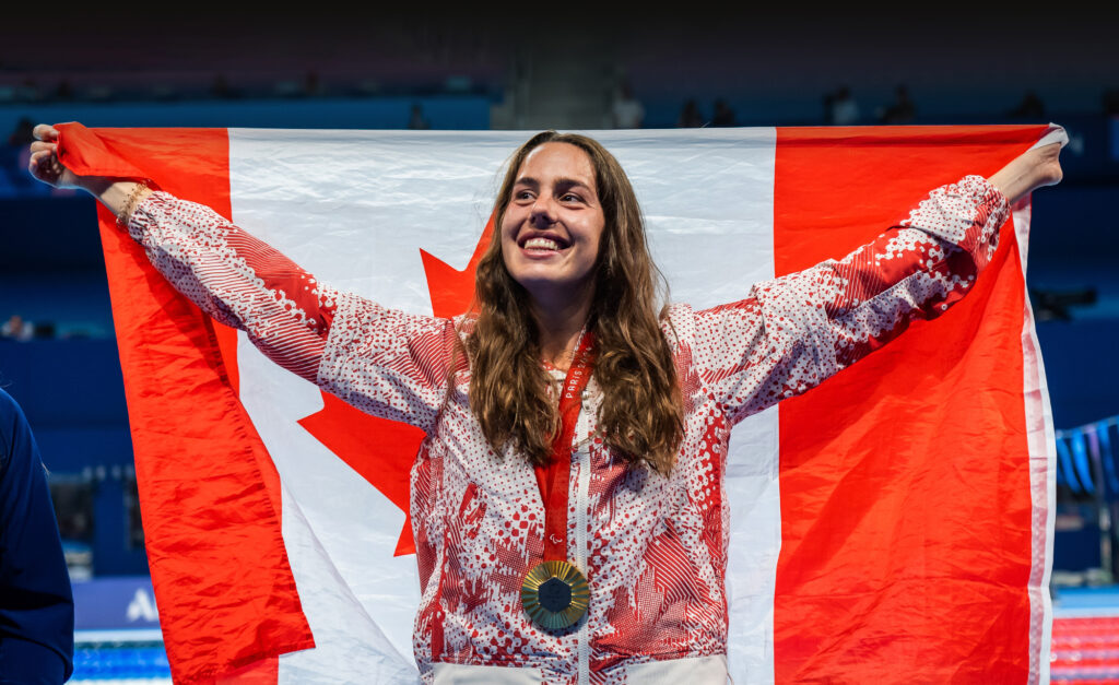 La nageuse paralympique canadienne Aurélie Rivard sourit fièrement en tenant un grand drapeau canadien derrière elle. Elle porte sa médaille de Paris 2024 et sa veste de podium d’Équipe Canada sur le bord de la piscine.
