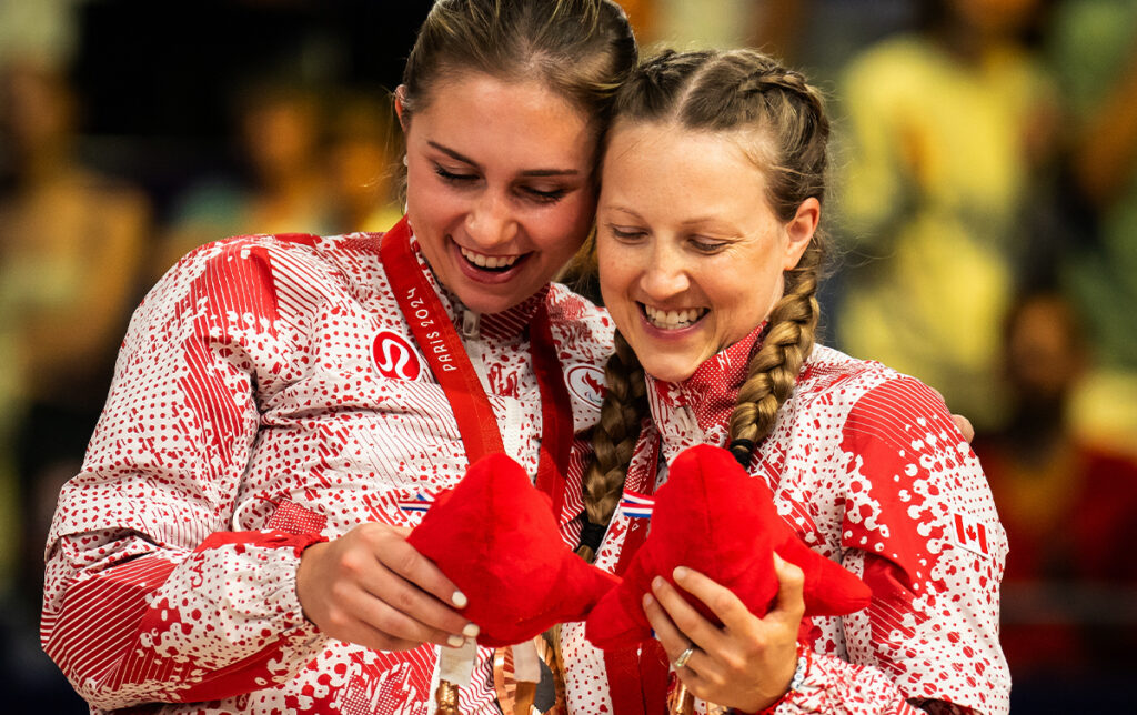 Two Canadian women’s sitting volleyball players smile and embrace while admiring their Paris 2024 bronze medals and plush mascots, wearing red and white Team Canada uniforms with braided hair and Lululemon logos visible.