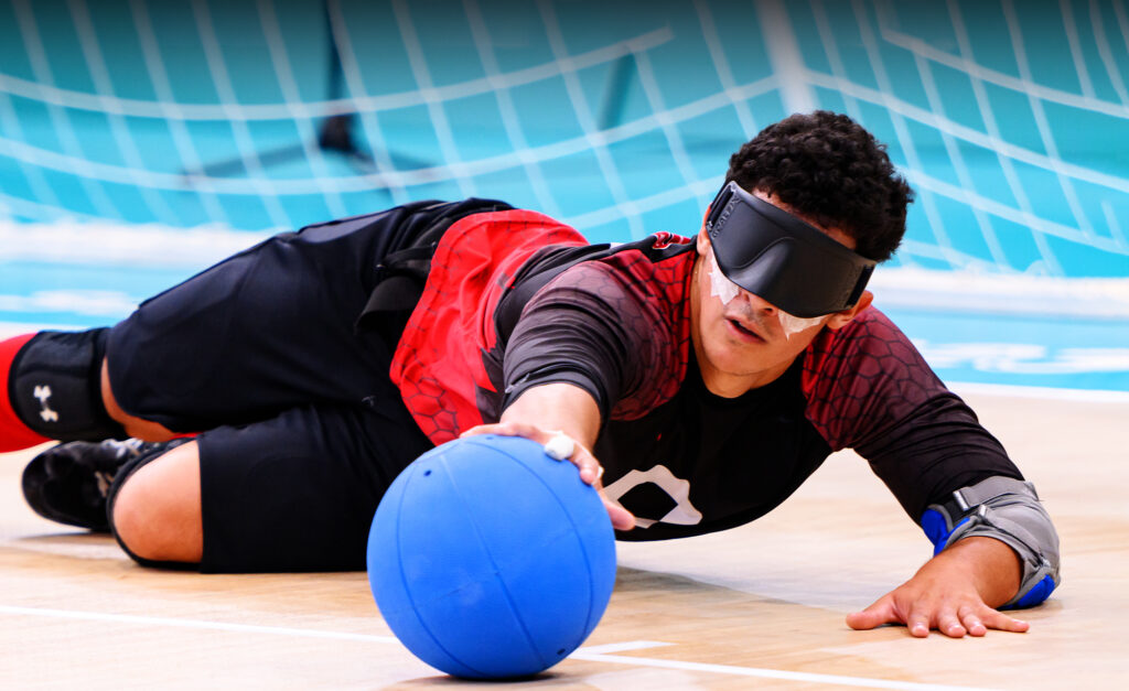 Un joueur canadien de goalball plonge pour bloquer un ballon pendant une compétition aux Jeux parapanaméricains de Santiago 2023, portant un masque oculaire et l’uniforme rouge et noir de l’équipe sur un terrain bleu clair.