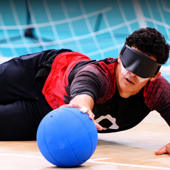 Un joueur canadien de goalball plonge pour bloquer un ballon pendant une compétition aux Jeux parapanaméricains de Santiago 2023, portant un masque oculaire et l’uniforme rouge et noir de l’équipe sur un terrain bleu clair.