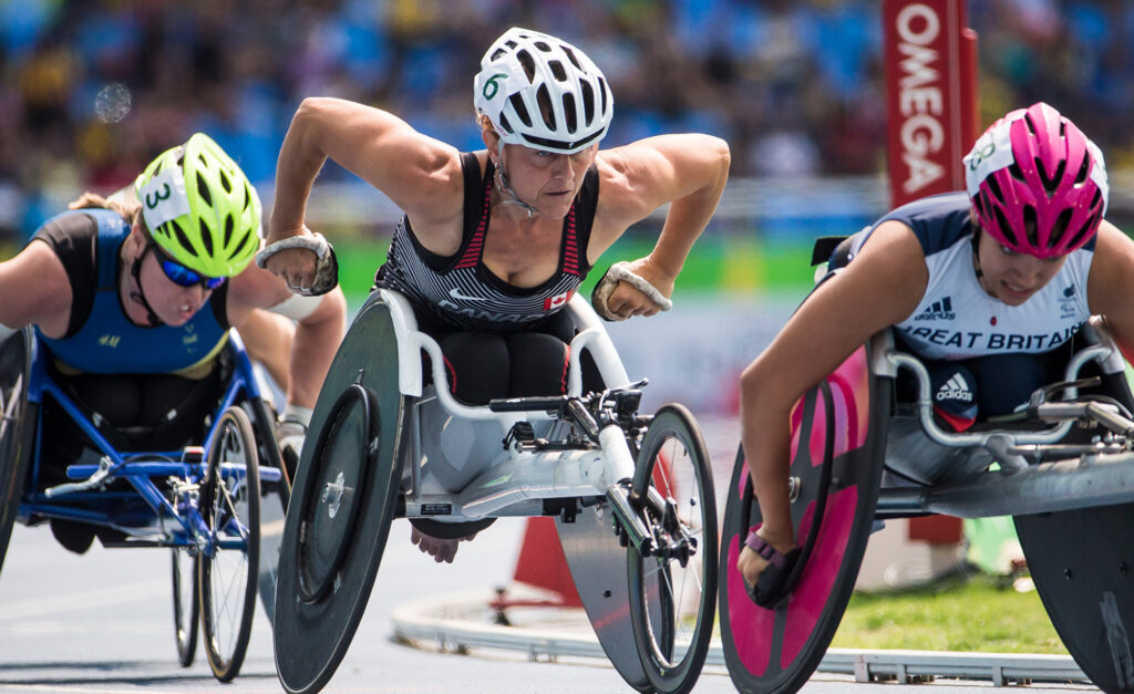 La paralympienne canadienne Diane Roy concourt avec intensité dans sa course en fauteuil roulant, sur une piste d’athlétisme, entre des athlètes de la Suède et de la Grande-Bretagne, affichant une concentration et une puissance remarquables.
