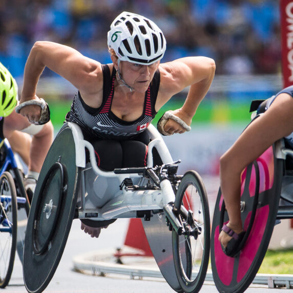 La paralympienne canadienne Diane Roy concourt avec intensité dans sa course en fauteuil roulant, sur une piste d’athlétisme, entre des athlètes de la Suède et de la Grande-Bretagne, affichant une concentration et une puissance remarquables.