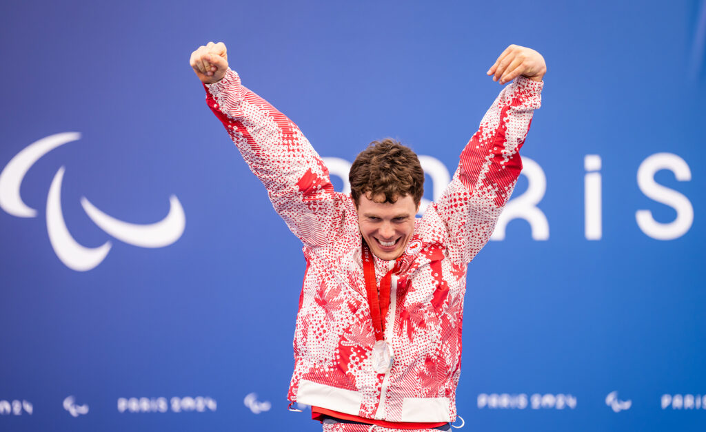 Le paranageur canadien Nathan Clement célèbre sur le podium avec les bras levés après avoir remporté une médaille d’argent aux Jeux paralympiques de Paris 2024. Il porte l’uniforme rouge et blanc d’Équipe Canada et sourit fièrement devant un fond bleu arborant le symbole paralympique et le mot « Paris ».