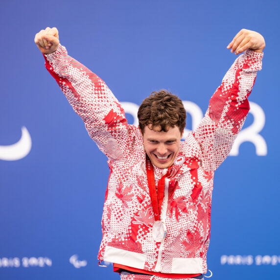 Le paranageur canadien Nathan Clement célèbre sur le podium avec les bras levés après avoir remporté une médaille d’argent aux Jeux paralympiques de Paris 2024. Il porte l’uniforme rouge et blanc d’Équipe Canada et sourit fièrement devant un fond bleu arborant le symbole paralympique et le mot « Paris ».
