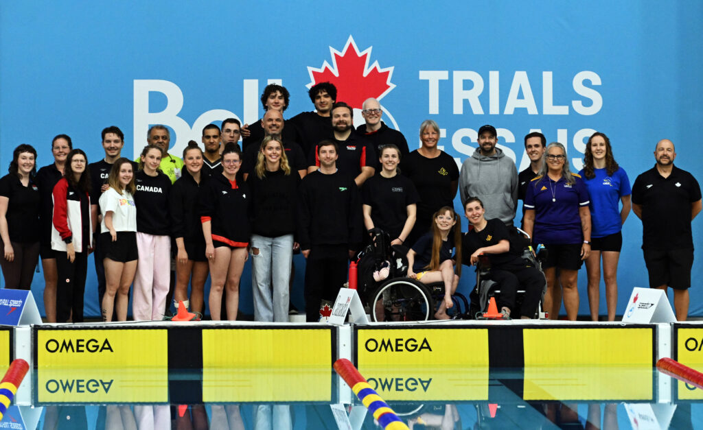 Photo de groupe de l’équipe paranatation et du personnel de Natation Canada sur le bord de la piscine devant une bannière bleue des « Essais Bell », avec les athlètes et entraîneurs souriants.