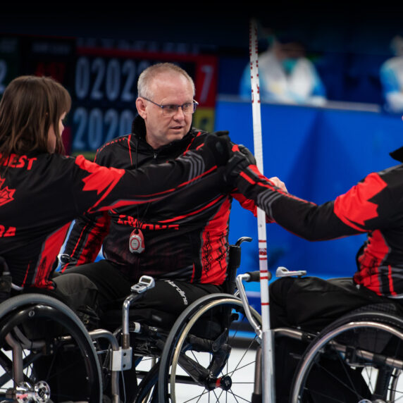 Les curleurs en fauteuil roulant canadiens Ina Forrest, Mark Ideson et Jon Thurston se donnent un coup de poing en guise d’encouragement lors d’un match aux Jeux paralympiques d’hiver de Beijing 2022. Les trois athlètes portent des vestes noires et rouges d’Équipe Canada avec leur nom inscrit au dos, assis dans leur fauteuil roulant sur la glace.