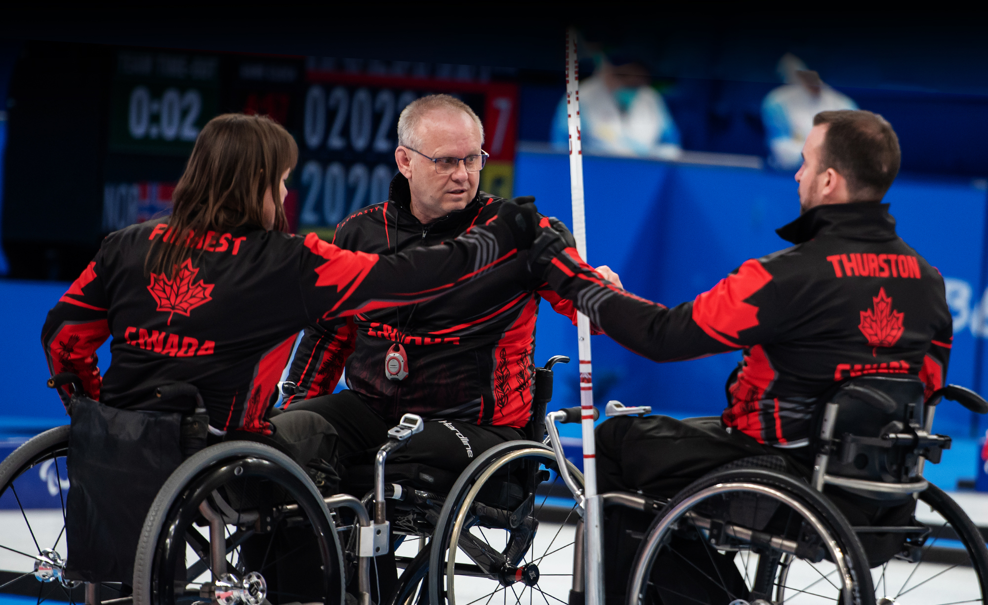 Les curleurs en fauteuil roulant canadiens Ina Forrest, Mark Ideson et Jon Thurston se donnent un coup de poing en guise d’encouragement lors d’un match aux Jeux paralympiques d’hiver de Beijing 2022. Les trois athlètes portent des vestes noires et rouges d’Équipe Canada avec leur nom inscrit au dos, assis dans leur fauteuil roulant sur la glace.