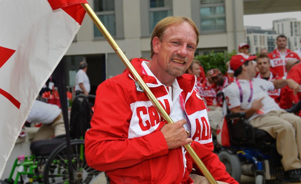Garett Hickling sourit en tenant fièrement le drapeau canadien, vêtu d’un manteau rouge et blanc d’Équipe Canada, entouré de ses coéquipiers lors d’un événement paralympique.