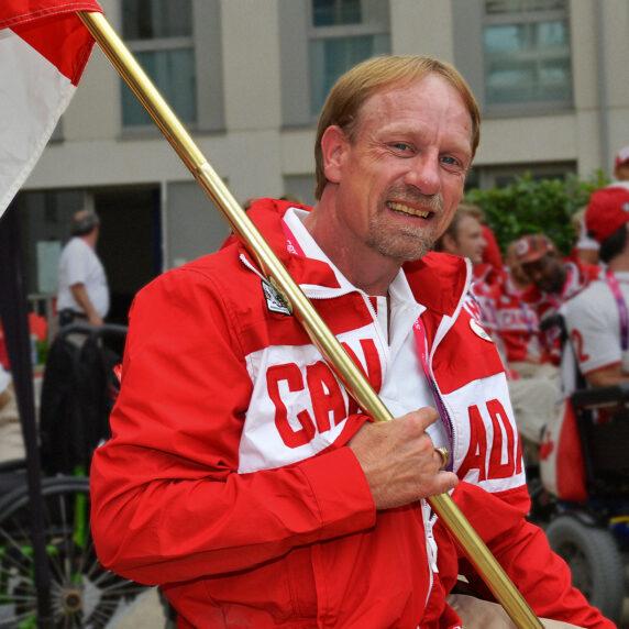 Garett Hickling sourit en tenant fièrement le drapeau canadien, vêtu d’un manteau rouge et blanc d’Équipe Canada, entouré de ses coéquipiers lors d’un événement paralympique.