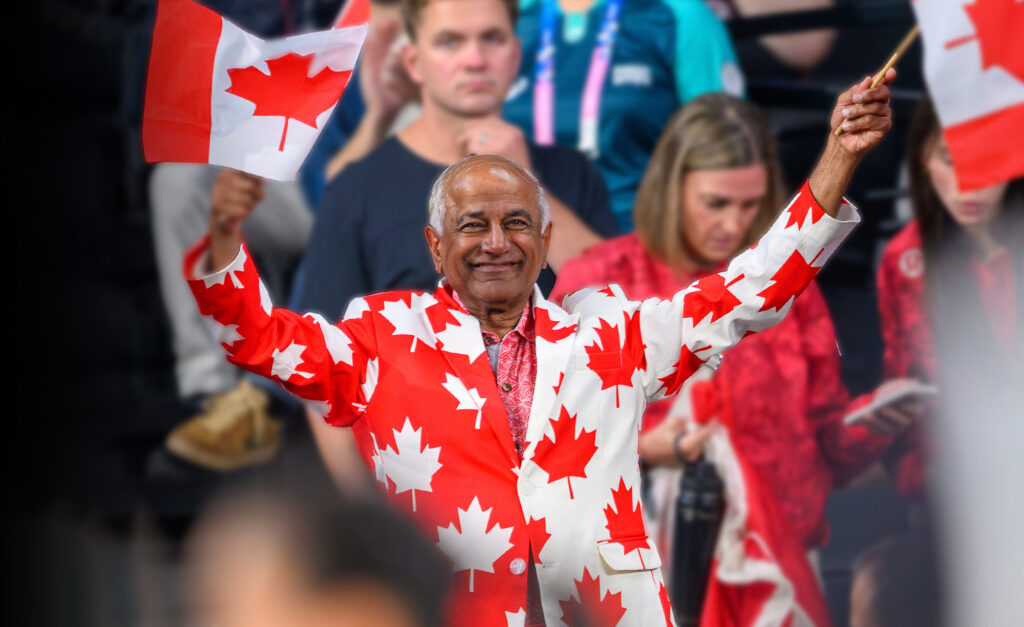 Mike Shaikh, membre du conseil d’administration de la Fondation paralympique canadienne, sourit et agite deux drapeaux canadiens en portant un habit orné de feuilles d’érable rouges lors d’un événement de l’Équipe paralympique canadienne.