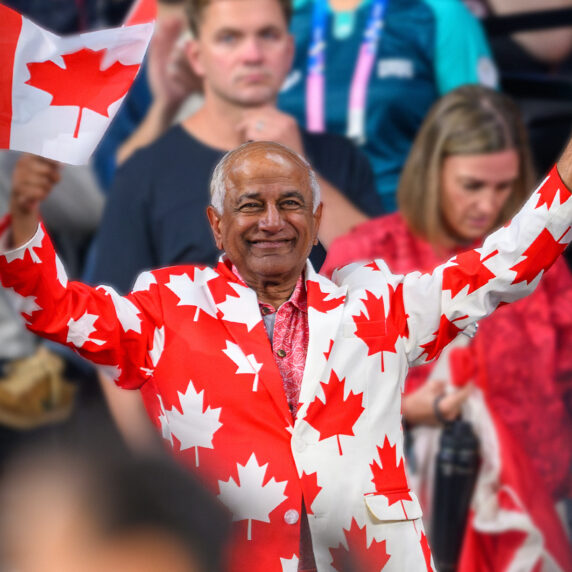 Mike Shaikh, membre du conseil d’administration de la Fondation paralympique canadienne, sourit et agite deux drapeaux canadiens en portant un habit orné de feuilles d’érable rouges lors d’un événement de l’Équipe paralympique canadienne.