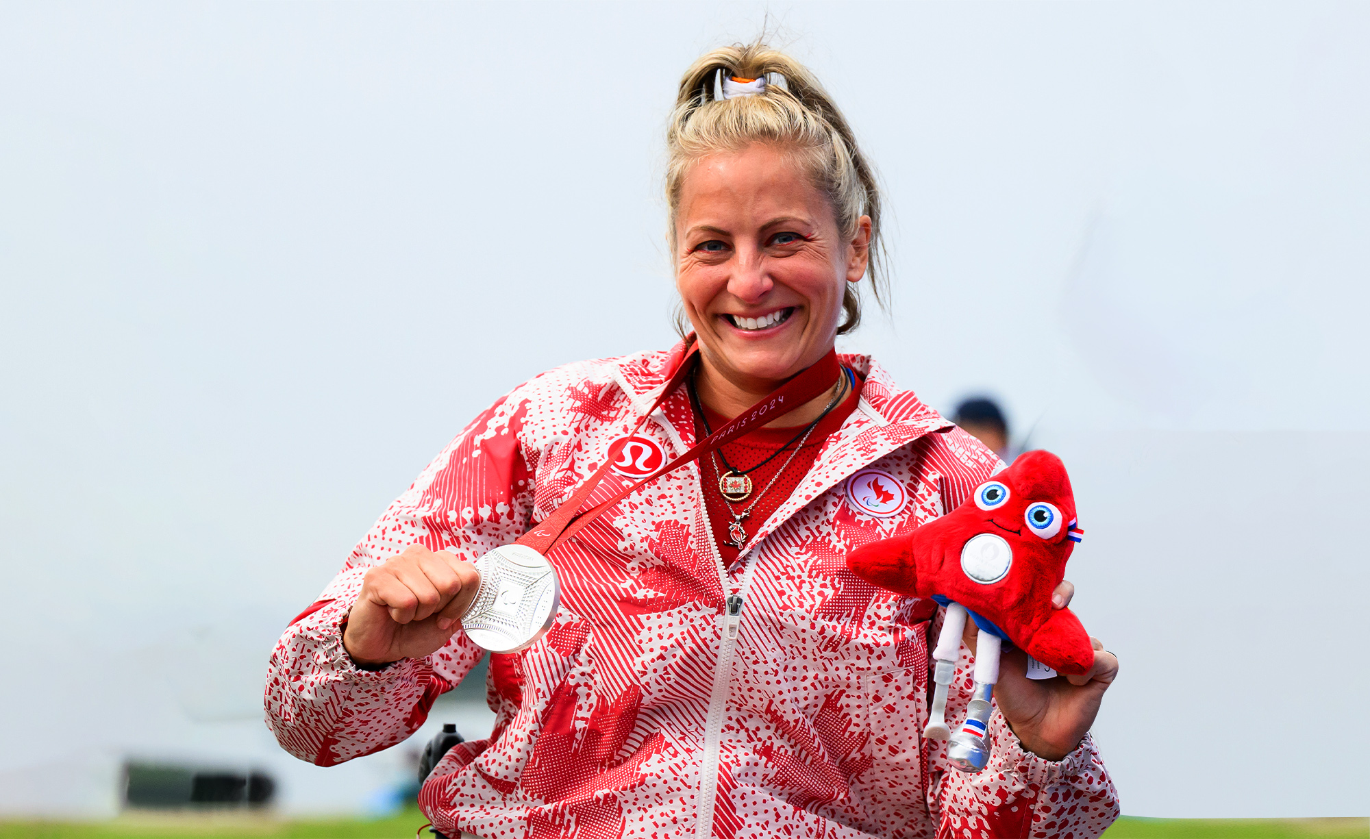Brianna Hennessy sourit fièrement en tenant sa médaille d’argent de Paris 2024 et la peluche rouge de la mascotte paralympique, vêtue de sa veste d’Équipe Canada ornée de feuilles d’érable.