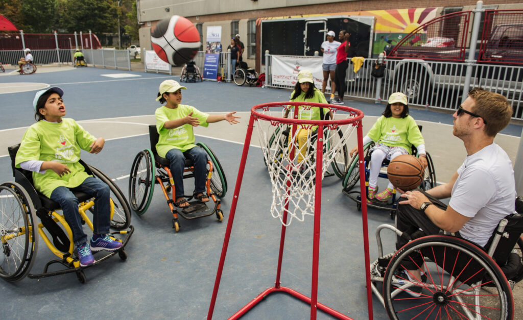Quatre enfants en fauteuils roulants et un adulte jouent au basketball en fauteuil roulant à l'extérieur, les enfants atteignant un ballon de basketball près d'un panier.
