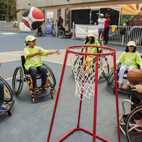 Quatre enfants en fauteuils roulants et un adulte jouent au basketball en fauteuil roulant à l'extérieur, les enfants atteignant un ballon de basketball près d'un panier.