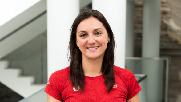 Portrait d’Alison Costa souriante, portant un chandail rouge d’Équipe Canada avec deux épinglettes du Comité paralympique canadien, devant un décor intérieur moderne aux tons blanc et gris.