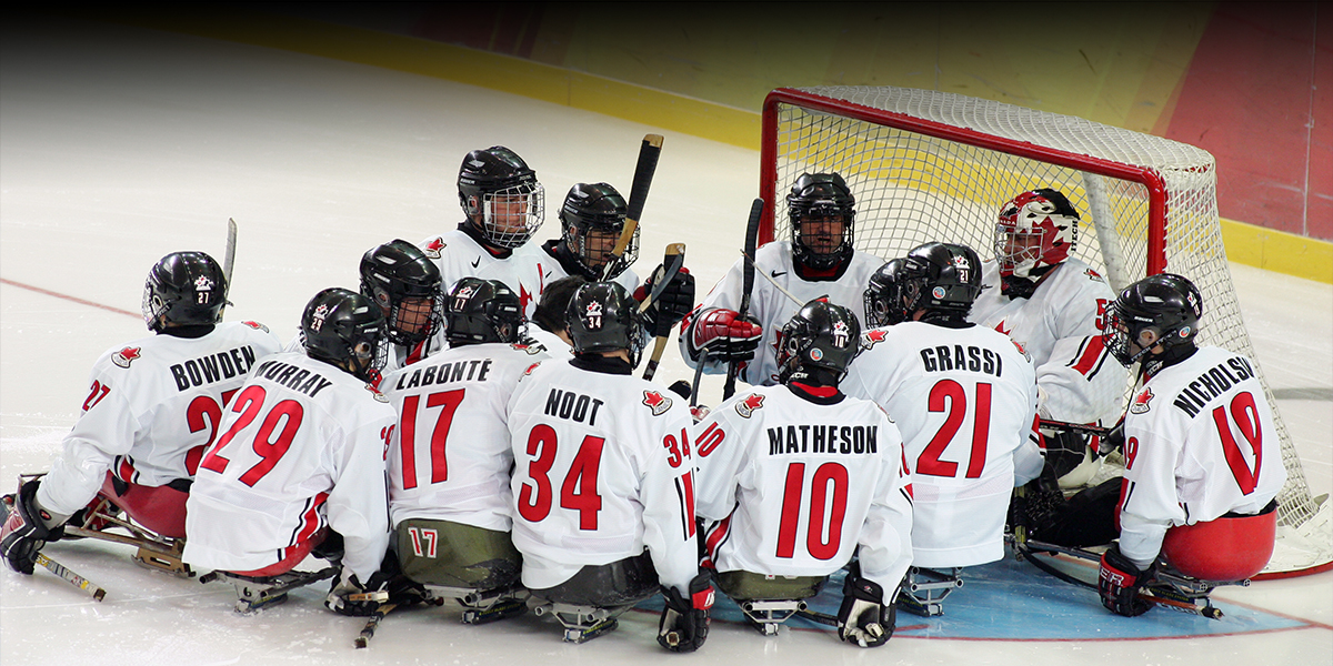 L’équipe canadienne de hockey sur luge réunie devant le but sur la glace aux Jeux paralympiques d’hiver de Torino 2006. Les joueurs, vêtus de chandails blancs avec des accents rouges, sont regroupés en cercle, certains tenant leur bâton verticalement.