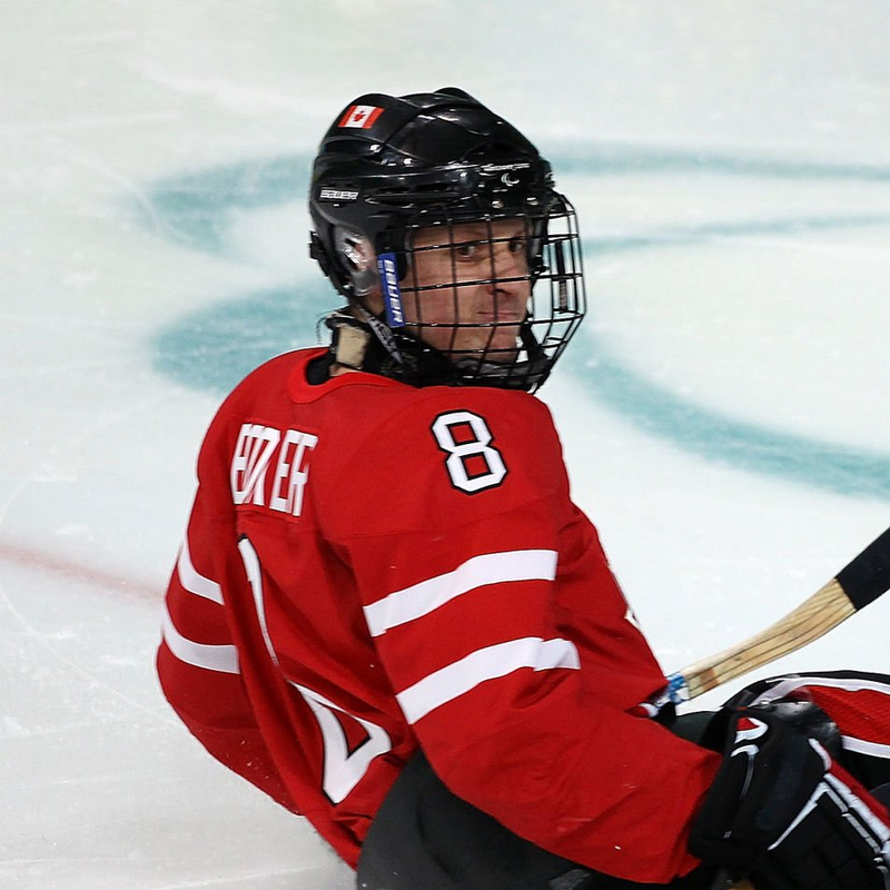 Portrait du joueur canadien de hockey sur luge Jeremy Booker, vêtu d’un chandail rouge portant le numéro 8, assis dans sa luge sur la glace pendant un match. Il porte un casque noir avec une grille complète et un autocollant du drapeau canadien sur le dessus.