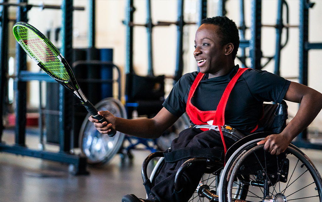 A young athlete in a wheelchair smiles while holding a tennis racquet, wearing a black shirt and red harness in an indoor gym setting.