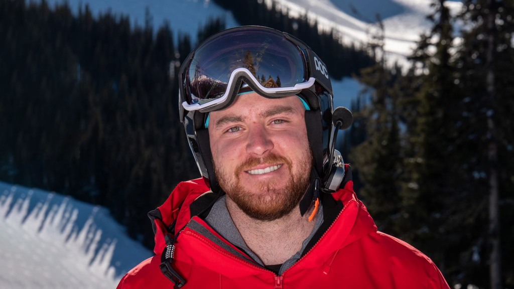Le paralympien canadien Mac Marcoux sourit en portant un casque de ski et une veste rouge, debout sur une pente enneigée avec des conifères en arrière-plan.