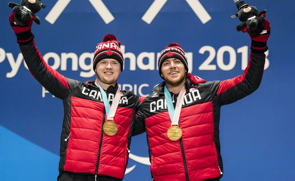 Le skieur para-alpin canadien Mac Marcoux et son guide célèbrent fièrement sur le podium aux Jeux paralympiques d’hiver de PyeongChang 2018, les bras levés, portant des médailles d’or ainsi que des manteaux et tuques d’Équipe Canada.