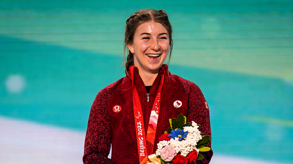 La skieuse alpine paranordique canadienne Mollie Jepsen sourit sur le podium des Jeux paralympiques d’hiver de Beijing 2022, portant sa veste rouge d’Équipe Canada, une médaille paralympique autour du cou et tenant un bouquet de fleurs de la victoire.