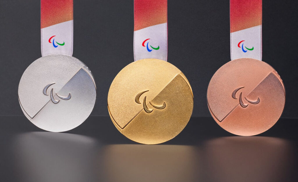Médailles d’or, d’argent et de bronze des Jeux paralympiques d’hiver de Milano Cortina 2026 exposées sur un fond foncé. Chaque médaille présente un design circulaire épuré avec une découpe diagonale et le symbole des agitos au centre, accompagnée d’un ruban rouge et blanc et d’une inscription en braille en relief.